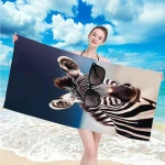Ręcznik plażowy 100x180 cm wykonany z mikrofibry, w kolorze czarno-białym z wizerunkiem zebry w okularach przeciwsłonecznych. Produkt trzymany przez modelkę na plaży, na tle morza i nieba z chmurami. Idealny do użytku na plaży i przy basenie.