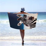 Ręcznik plażowy 100x180 cm w kolorach czerni i bieli z wzorem zebry i okularami przeciwsłonecznymi. Ułożony jest poziomo, trzymany przez osobę stojącą nad brzegiem morza. Tło przedstawia plażę i wodę, co podkreśla jego plenerowy charakter.