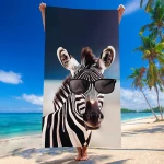 Duży ręcznik plażowy w rozmiarze 100x180 cm, wykonany z mikrofibry, z kolorowym wzorem przedstawiającym twarz zebry w okularach przeciwsłonecznych. Produkt trzymany jest w pozycji pionowej na plaży, w tle widać palmę i morze.
