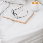Białe prześcieradło bawełniane hotelowe 140x200 leży na łóżku, obok otwarta książka i filiżanka kawy. Okulary w czarnej oprawie znajdują się na stronach książki. Tło jest jasne, a wszystkie elementy są dobrze widoczne.