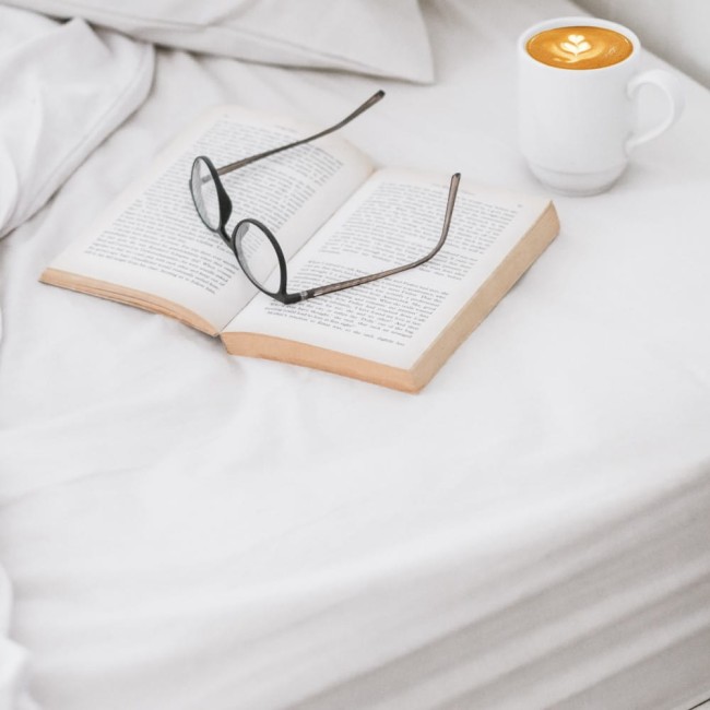 Prześcieradło bawełniane hotelowe białe 220x240, gładkie, leżące na łóżku. W tle otwarta książka z okrągłymi okularami oraz filiżanka kawy z wzorem na powierzchni. Tło neutralne, białe, co podkreśla produkt.