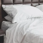 Białe, gładkie prześcieradło bawełniane hotelowe o wymiarach 220x240 cm, leżące na łóżku. Widoczne poduszki z poszewkami w tym samym kolorze. Tło stanowi ciemna, pikowana tapeta. Tekstura materiału sprawia wrażenie miękkości i gładkości.