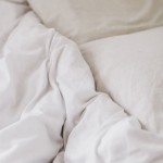 Prześcieradło bawełniane hotelowe białe 160x240, gładkie, leżące na łóżku obok dwóch poduszek. Widoczna faktura materiału, utrzymane w neutralnych kolorach. Ujęcie z góry, ukazujące rozłożenie prześcieradła na jasnym tle.