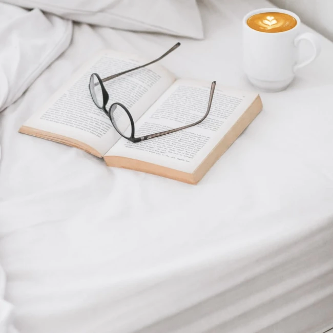 Prześcieradło bawełniane hotelowe, białe, o wymiarach 140x260 cm, widoczne na rozłożonym łóżku, dominuje w tle. Ułożone na białej pościeli, obok znajduje się otwarta książka i filiżanka kawy, tworząc przytulną atmosferę.