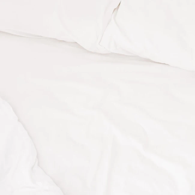 Prześcieradło bawełniane hotelowe białe 140x260, gładkie, leżące na łóżku. Tło jednolite, jasne, podkreślające kolor prześcieradła. Widoczne także poduszki w tym samym kolorze, tworzące spójną aranżację.
