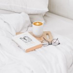 Białe, gładkie prześcieradło bawełniane rozłożone na łóżku. Na nim leży drewniana taca z kubkiem kawy oraz książka z białą okładką. Szklane okulary w metalowej oprawie są obok tacy. Tło miękkie, w odcieniach bieli i szarości.