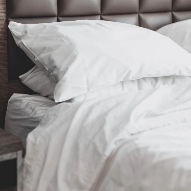 Prześcieradło bawełniane hotelowe białe 140x260, gładkie, leży na łóżku z poduchami. Ujęcie z boku, na tle ciemnego zagłówka. Widać delikatne zmarszczenia materiału, co podkreśla jego miękkość i fakturę.