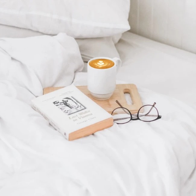 Białe, gładkie prześcieradło bawełniane o wymiarach 140x220 cm leży na łóżku. Obok znajduje się drewniana deska z filiżanką kawy oraz książką „Last Waltz in Vienna”, a także okrągłe okulary. Tło jest jasne i stonowane.