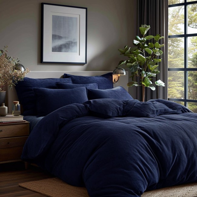 Poszewka frotte 40x40 w ciemnym granacie, leżąca na łóżku w aranżacji sypialni. Widoczne różne rozmiary i kształty poduszek w tym samym kolorze na tle jasnej ściany. W tle lampa oraz roślina doniczkowa przy oknie.