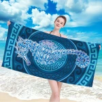Ręcznik plażowy 100x180 cm w kolorze błękitnym z naszytym wizerunkiem konika morskiego na ciemnym tle. Produkt rozłożony, trzymany przez kobietę na tle plaży. Wzorowany na greckich ornamentach, wykonany z mikrofibry, szybko schnie.