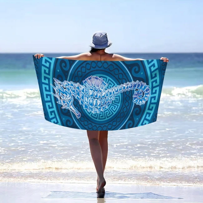 Ręcznik plażowy 100x180 cm z mikrofibry z grafiką konika morskiego. Kolory błękitny i turkusowy. Produkt rozłożony, trzymany przez osobę stojącą na plaży, w tle morze. Szybkoschnący materiał idealny na plażę.