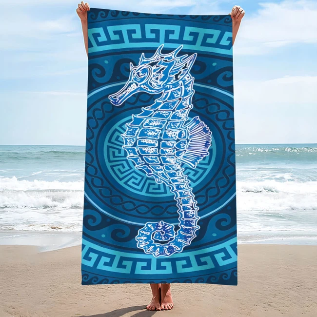 Ręcznik plażowy 100x180 cm wykonany z mikrofibry, z motywem konika morskiego w odcieniach błękitu, na wzorzystym tle. Produkt trzymany w pozycji stojącej nad brzegiem morza, widoczny detal graficzny konika morskiego oraz okrągły wzór dekoracyjny.