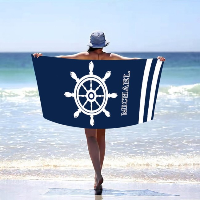 Ręcznik plażowy 100x180 cm w kolorze granatowym z białym wzorem przedstawiającym koło ratunkowe i napisem MICHAEL. Ujęcie z tyłu, osoba trzyma ręcznik rozłożony nad morzem. Widoczne tekstura mikrofibry, plażowe otoczenie.
