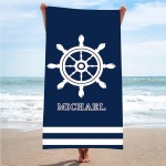 Ręcznik plażowy 100x180 cm w kolorze granatowym z białymi paskami u dołu. Na głównym tle znajduje się wzór w postaci steru oraz napis MICHAEL. Ręcznik trzymany jest przez osobę na plaży z widocznym tłem morza i nieba.