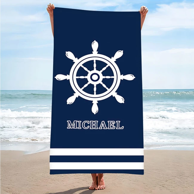 Ręcznik plażowy 100x180 cm w kolorze granatowym z białymi paskami u dołu. Na głównym tle znajduje się wzór w postaci steru oraz napis MICHAEL. Ręcznik trzymany jest przez osobę na plaży z widocznym tłem morza i nieba.