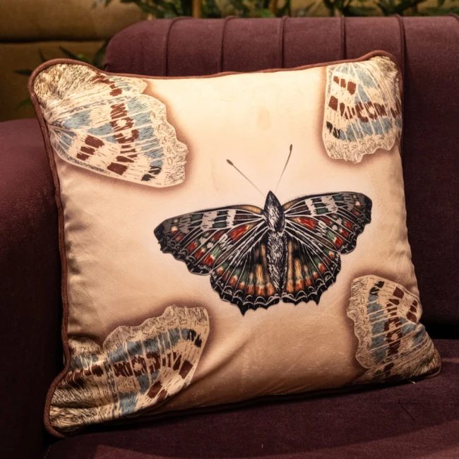 Poszewka dekoracyjna 43x43 cm w kremowym kolorze, z dużym, kolorowym motylem na środku oraz mniejszymi motylami w rogach. Produkt leży płasko, na tle ciemnofioletowej kanapy, z subtelnym połyskiem materiału.