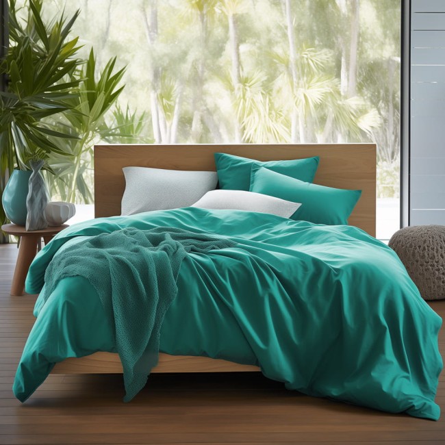 Poszewka satynowa 50x70 w kolorze ciemnego turkusu leży na łóżku, otoczona poduszkami w różnych odcieniach turkusu i bieli. W tle widoczna jest jasna przestrzeń z dużymi oknami i roślinnością.