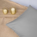 Poszewka satynowa 50x70 w kolorze popielatym leży na beżowym tle. Widoczna jest gładka faktura materiału. W pobliżu znajdują się dwa świeczniki w złotych osłonkach z białymi świecami. Ujęcie z boku podkreśla elegancki wygląd poszewki.