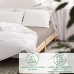 Prześcieradło satynowe z gumką 120x200 w kolorze ciemnego beżu leży na łóżku z jasną, dekoracyjną pościelą. Ujęcie z boku pokazuje tkaninę o gładkiej, błyszczącej fakturze. Widoczne są dwa białe poduszki na tle. Tło stanowi jasne wnętrze z zasłonami.