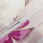 Zbliżenie na zamek błyskawiczny pościeli satynowej w kolorze białym z różowymi kwiatami. Widoczne detale wzoru kwiatowego oraz gładka faktura materiału. Zamek wykonany w jasnym kolorze, zamknięty, na tle jasnej tkaniny.