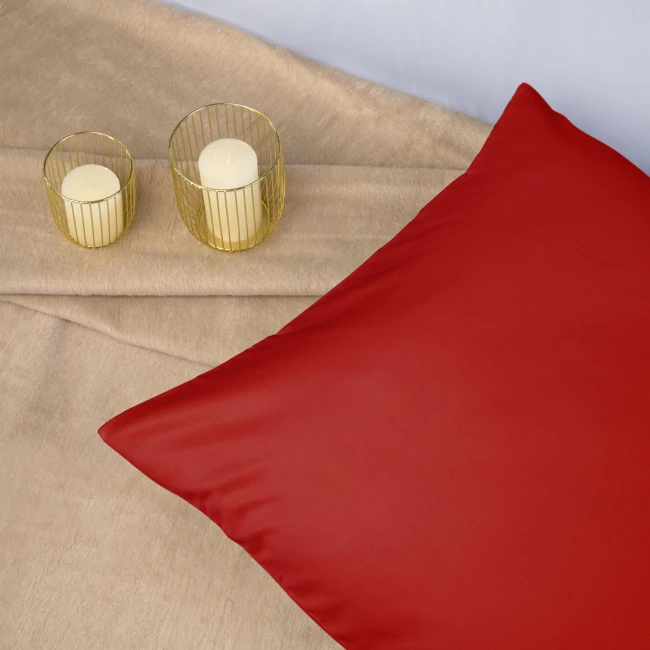 Poszewka satynowa 50x70 w kolorze intensywnej czerwieni, leżąca na beżowym tle, z delikatnym połyskiem. Obok znajdują się dwie świeczki w złotych świecznikach. Kąt ujęcia z góry, pokazujący całość produktu oraz jego otoczenie.