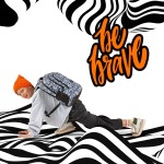 Plecak szkolny BackUp w czarno-białym wzorze, z niebieskimi akcentami. W ujęciu ukośnym, na modelu w pozycji klęczącej. Tło z graficznymi falami, widoczne logo na plecaku oraz napis be brave w pomarańczowym kolorze.