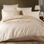 Pościel satyna bawełniana żakardowa 160x200 w odcieniu jasnego beżu, z subtelnym wzorem na poszewkach i narzucie. Złożona na łóżku z poduszkami w różnych rozmiarach, na tle ciemnej ściany i lampy stołowej. Widoczne detale tkaniny i struktury.