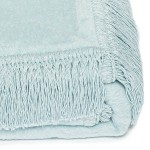 Koc bawełniany 150x200 w jasnym niebieskim kolorze, z frędzlami na krótszym brzegu. Widok z boku, koc leży zwinięty, ukazując fakturę materiału i delikatny odcień błękitu. Tło białe, co podkreśla kolor i detale zabezpieczeń brzegów.