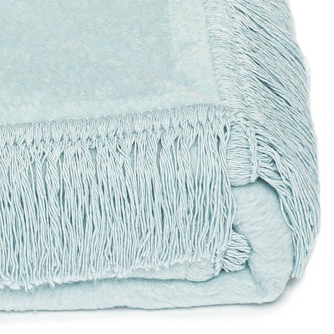 Koc bawełniany 150x200 w jasnym niebieskim kolorze, z frędzlami na krótszym brzegu. Widok z boku, koc leży zwinięty, ukazując fakturę materiału i delikatny odcień błękitu. Tło białe, co podkreśla kolor i detale zabezpieczeń brzegów.