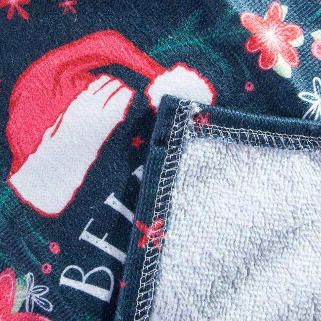 Ścierka kuchenna w rozmiarze 38x60 cm, z nadrukiem Świętego Mikołaja oraz reniferów na ciemnym tle. Materiał miękki z teksturą, boki wykończone ozdobnym ściegiem. Złożona w połowie, prezentująca detale wzoru i faktury.