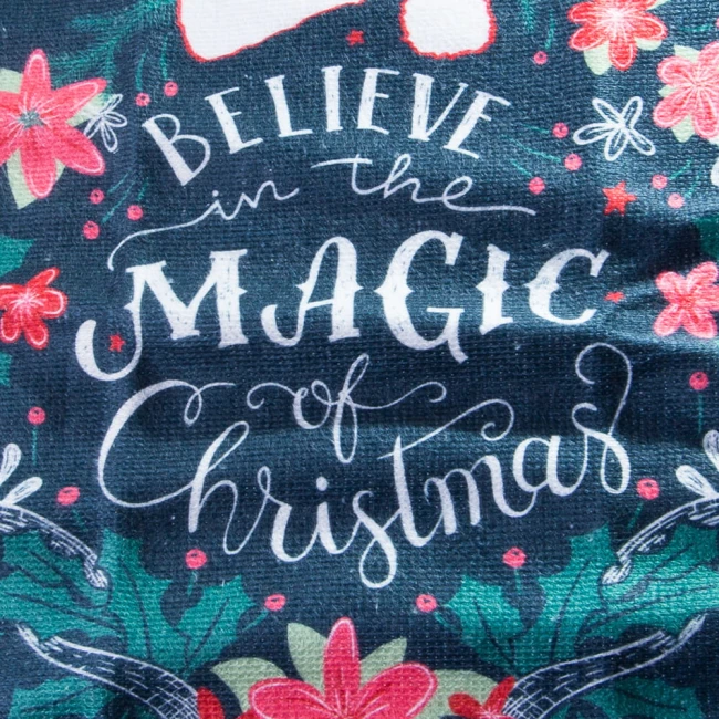 Ścierka kuchenna w kolorze granatowym, z widocznym napisem BELIEVE in the MAGIC of Christmas oraz motywami roślinnymi, takimi jak czerwone kwiaty i zielone liście. Zbliżenie na materiał z fakturą, bez zbędnych dekoracji.
