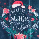 Ścierka kuchenna w ciemnoniebieskim kolorze z nadrukiem świątecznym. Na tkaninie widoczny tekst „Believe in the Magic of Christmas” otoczony kwiatami w odcieniach różu i zieleni, z białymi akcentami. Produkt leży na płaskiej powierzchni.