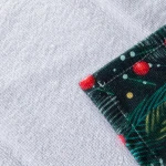 Ścierka kuchenna z motywem świątecznym, biało-zielona, grodzona z czerwonymi elementami. Widoczne są detale roślinne i krople dekoracyjne. Złożona w rogu, prezentuje teksturę materiału. Wymiary 38x60 cm, idealna do dekoracji podczas gotowania.