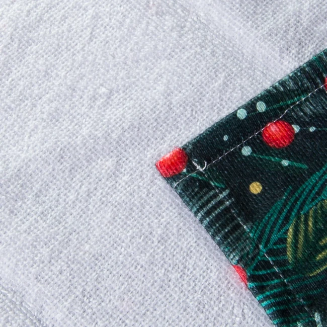 Ścierka kuchenna z motywem świątecznym, biało-zielona, grodzona z czerwonymi elementami. Widoczne są detale roślinne i krople dekoracyjne. Złożona w rogu, prezentuje teksturę materiału. Wymiary 38x60 cm, idealna do dekoracji podczas gotowania.