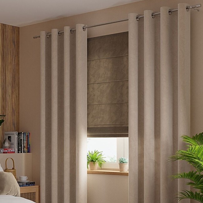 Zasłona gotowa welwetowa 140x260 cm w ciemno beżowym kolorze, wisząca na przelotkach. Gładka, miękka tkanina z teksturą. Ujęcie frontalne, w otoczeniu minimalistycznej aranżacji wnętrza z roślinami i książkami w tle.