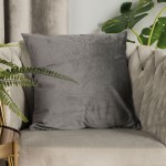 Poszewka welwetowa 40x40 w ciemno szarej kolorystyce, o gładkiej fakturze, leży na jasnym, eleganckim fotelu. Tło w kremowym odcieniu, otoczenie ozdobione zielonymi liśćmi roślin. Produkt idealny do dekoracji wnętrz.