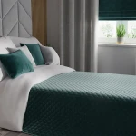 Poszewka welwetowa 40x40, ciemno turkusowa, umieszczona na białej pościeli. Widoczne są też dwa poduszki w odcieniach turkusu oraz szarości. Zdjęcie z perspektywy z boku, w jasno urządzonym wnętrzu z rośliną w doniczce obok okna.