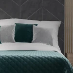 Poszewka welwetowa 40x40 w kolorze ciemno turkusowym, umieszczona na łóżku z białą pościelą. W tle poduszki w odcieniach szarego i turkusowego. Tekstura welwetu widoczna w zbliżeniu na kolorową powierzchnię. Produkt leży na miękkim materiale.