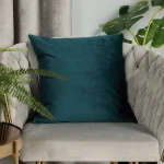 Poszewka welwetowa 40x40, ciemno turkusowa, na prostokątnej poduszce, leżąca na eleganckim, jasnoszarym fotelu. Otoczona rośliną doniczkową. Gładka, teksturowana powierzchnia, brak widocznych napisów czy logo.