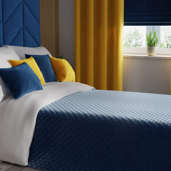 Poszewka welwetowa 40x40 w ciemnoniebieskim kolorze, z teksturą welwetu, leży na białej kołdrze. W tle widoczne są żółte zasłony oraz okno z roletą. Poduszki w różnych kolorach (ciemnoniebieska i żółta) układają się na głowie łóżka.