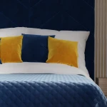 Ciemno niebieska poszewka dekoracyjna 40x40 wykonana z welwetu, leżąca na białej pościeli. W tle widnieją dwie żółte i jedna niebieska poduszka. Produkt w aranżacji sypialni, na tle strukturalnej ściany w odcieniach granatu.