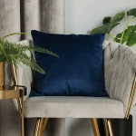 Poszewka welwetowa 40x40 w ciemno niebieskim kolorze, umieszczona na szarej, miękkiej kanapie. Produkt jest rozłożony, z widoczną, gładką fakturą. W tle roślinność oraz złote akcenty mebli.