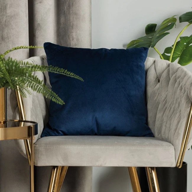 Poszewka welwetowa 40x40 w ciemno niebieskim kolorze, umieszczona na szarej, miękkiej kanapie. Produkt jest rozłożony, z widoczną, gładką fakturą. W tle roślinność oraz złote akcenty mebli.