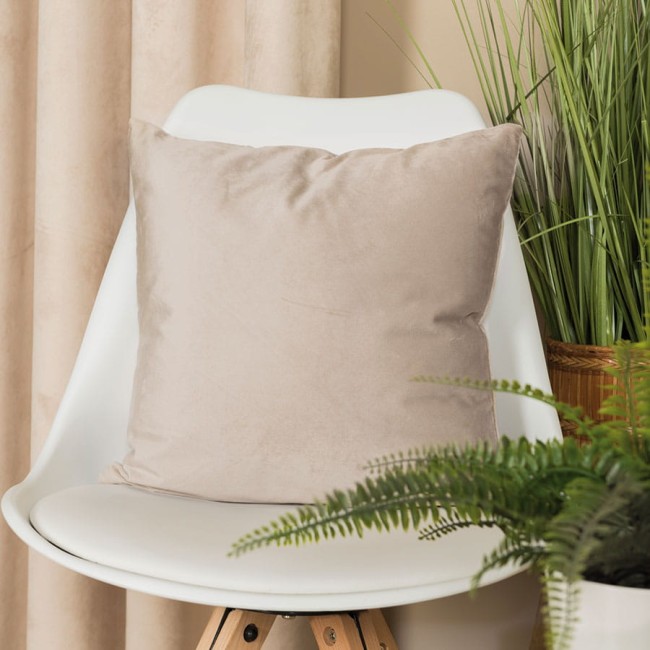 Poszewka welwetowa 40x40, ciemno kremowa, umieszczona na białym krześle. Produkt leży na siedzisku, ukazując miękką, teksturowaną fakturę materiału. W tle widoczne są rośliny oraz zasłony w stonowanych barwach, co tworzy przytulną atmosferę.