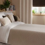 Poszewka welwetowa 40x40 w kremowym kolorze leży na białej pościeli na łóżku, z innymi poduszkami w odcieniach beżu i oliwkowej zieleni. Tło w jasnych tonacjach z zasłonami i roślinami w doniczkach. Detaile poszewki są widoczne w zbliżeniu.