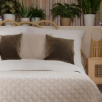 Poszewka welwetowa 40x40 Velvi w kolorze kremowym, z delikatną fakturą, leży na łóżku. Na poduszce znajdują się trzy dekoracyjne poduszki: dwie w odcieniu brązowym i jedna w jasnym kolorze. W tle rośliny doniczkowe i naturalne materiały.