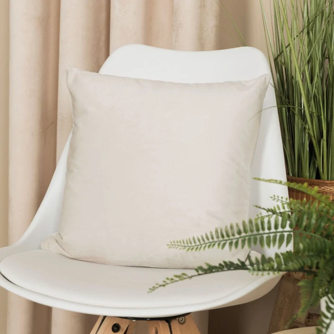 Poszewka dekoracyjna welwetowa 40x40 w kolorze kremowym, umieszczona na białym krześle. Pozycja leżąca, z bliskim ujęciem, ukazującym tkaninę i fakturę welwetu. W tle widoczne zasłony i roślinność, co tworzy przytulny klimat aranżacyjny.