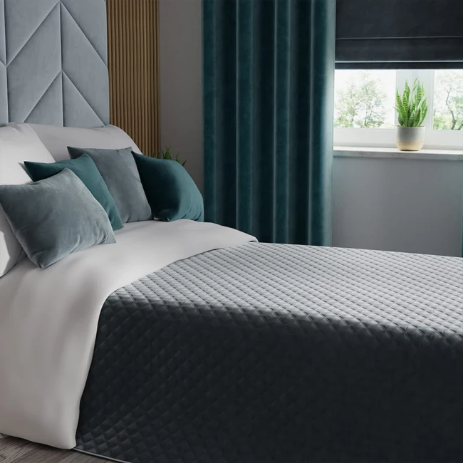 Poszewka welwetowa 40x40 w kolorze niebieskim na szarym łóżku. Ułożona leżąco, z dekoracyjnymi poduszkami w odcieniach szarości i zieleni. Tło z zasłonami z welwetu, oknem oraz rośliną doniczkową. Wzór przeszycia na tkaninie widoczny na pościeli.