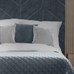 Poszewka welwetowa 40x40 Velvi w kolorze niebieskim, umieszczona na białej pościeli z dwoma dekoracyjnymi poduszkami w odcieniach szarości. Ujęcie frontalne, detal materiału widoczny. Tło w stonowanych kolorach, z elementami drewnianymi.