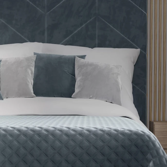 Poszewka welwetowa 40x40 Velvi w kolorze niebieskim, umieszczona na białej pościeli z dwoma dekoracyjnymi poduszkami w odcieniach szarości. Ujęcie frontalne, detal materiału widoczny. Tło w stonowanych kolorach, z elementami drewnianymi.